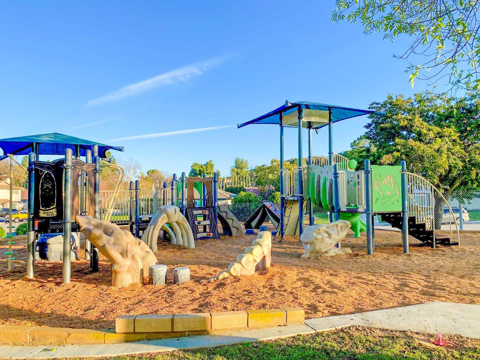 A colorful playground with slides, climbing structures, and wood chips on the ground, set in a park with trees and a clear sky.