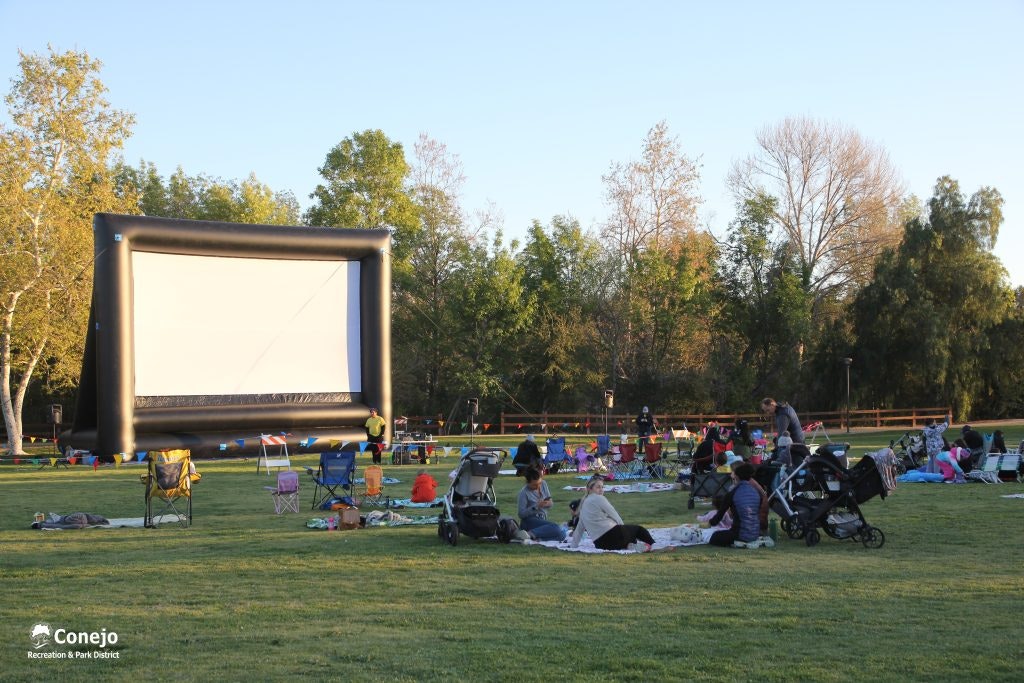 People seated on blankets and chairs in a park, facing a large inflatable screen, ready for an outdoor movie event.