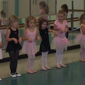 Young girls in ballet attire standing in a row, practicing in a dance studio with mirrors.