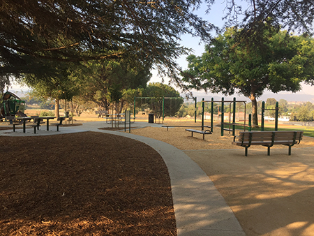 Outdoor playground with benches, swing sets, trees, and walking paths under clear skies.