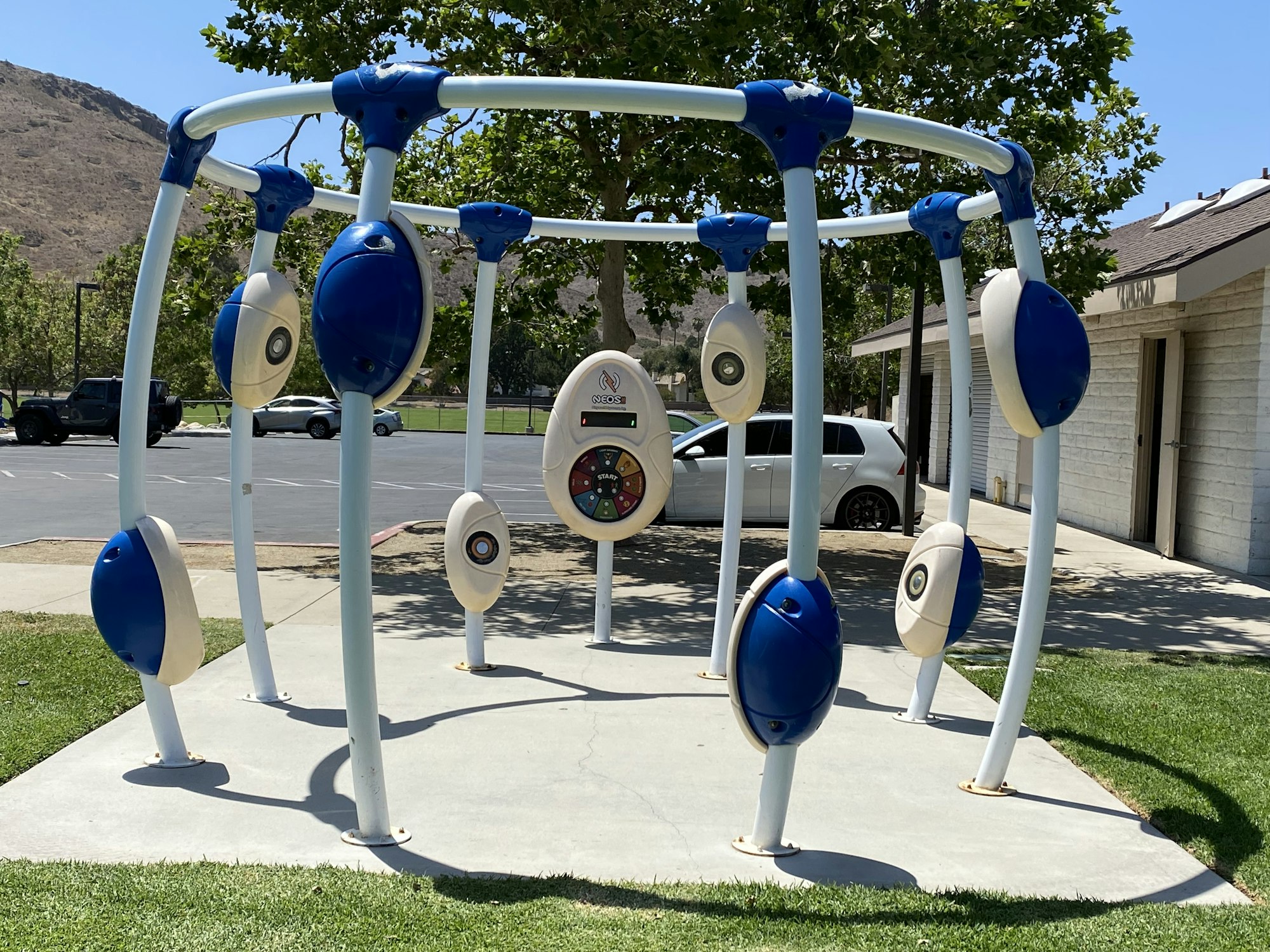 A musical playground structure with various blue and white panels, set on concrete with trees and parked cars nearby.