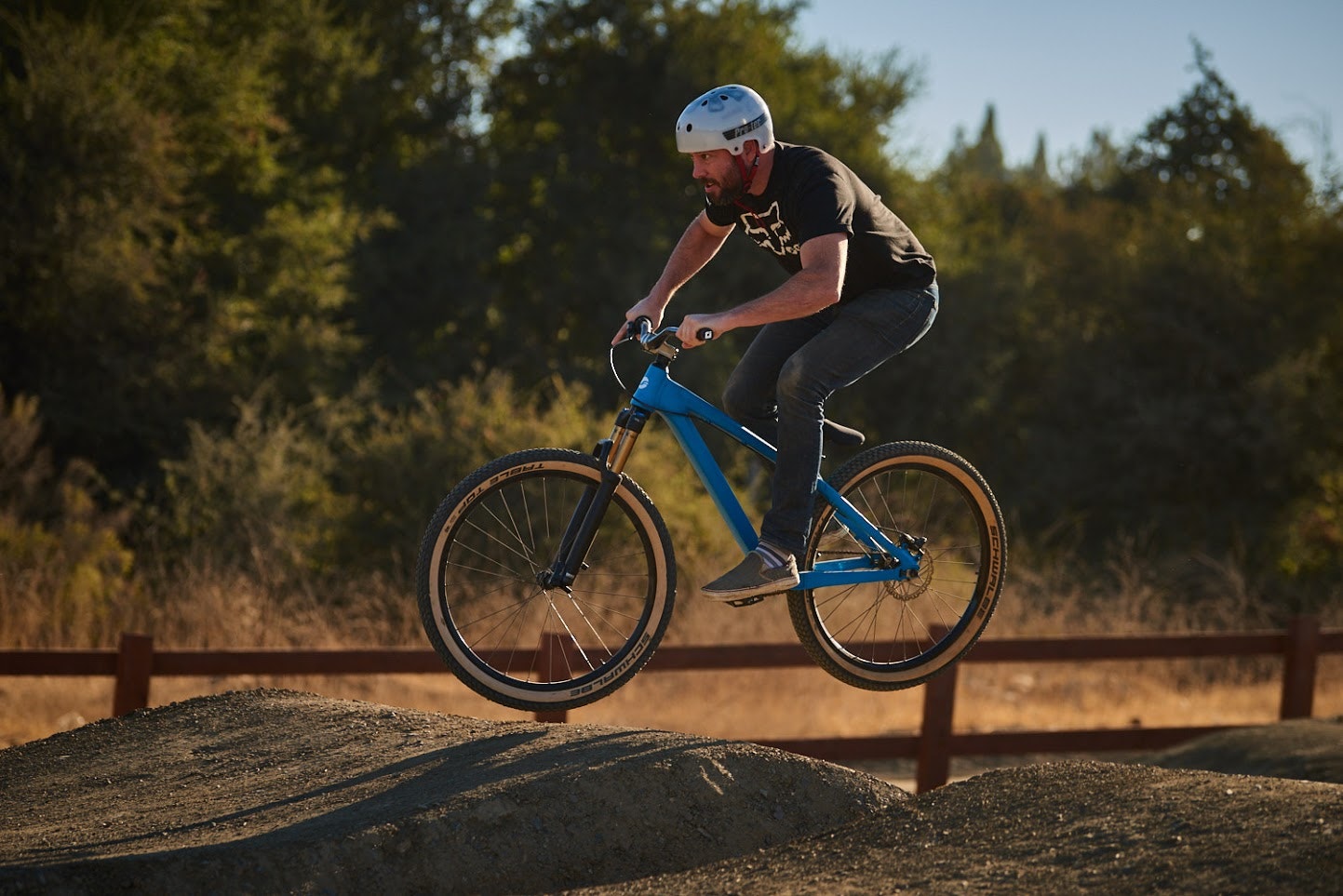 A person wearing a helmet is riding a blue mountain bike, mid-air, on a dirt trail with trees in the background.