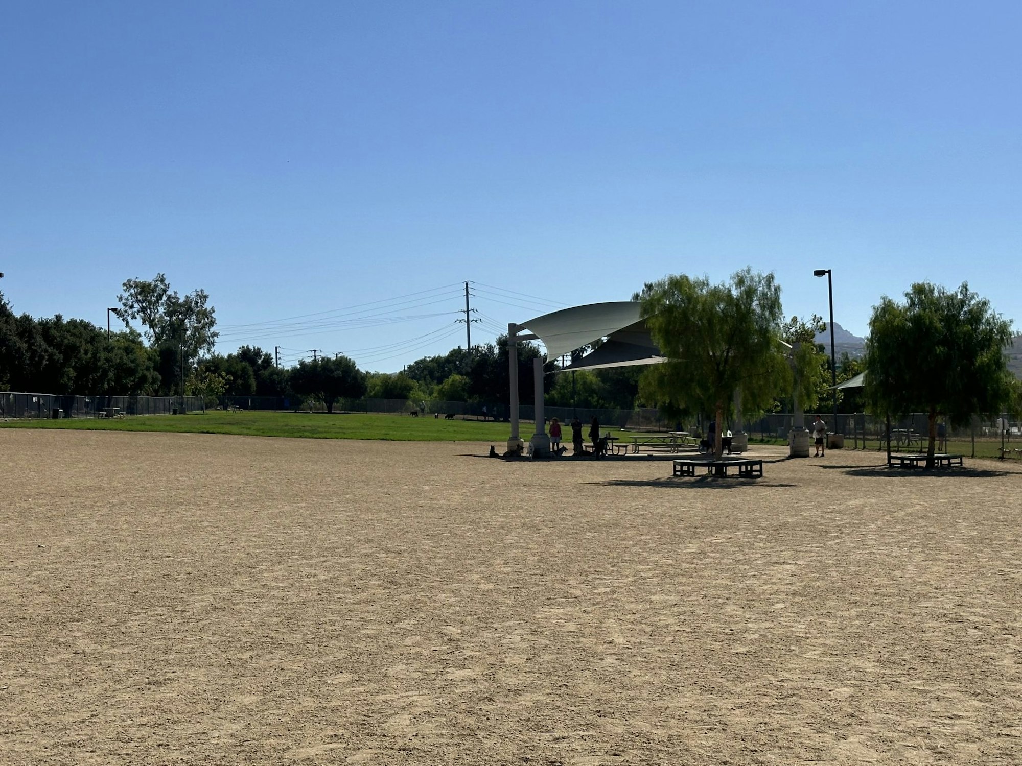 A park with a large sandy area, trees, picnic tables, and people in the shade.