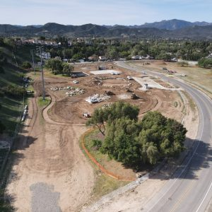 An aerial view of a construction site with heavy machinery, dirt roads, and nearby greenery, surrounded by a curving road.