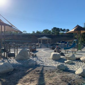 A playground with slides, rocks, and shaded structures under a clear blue sky.