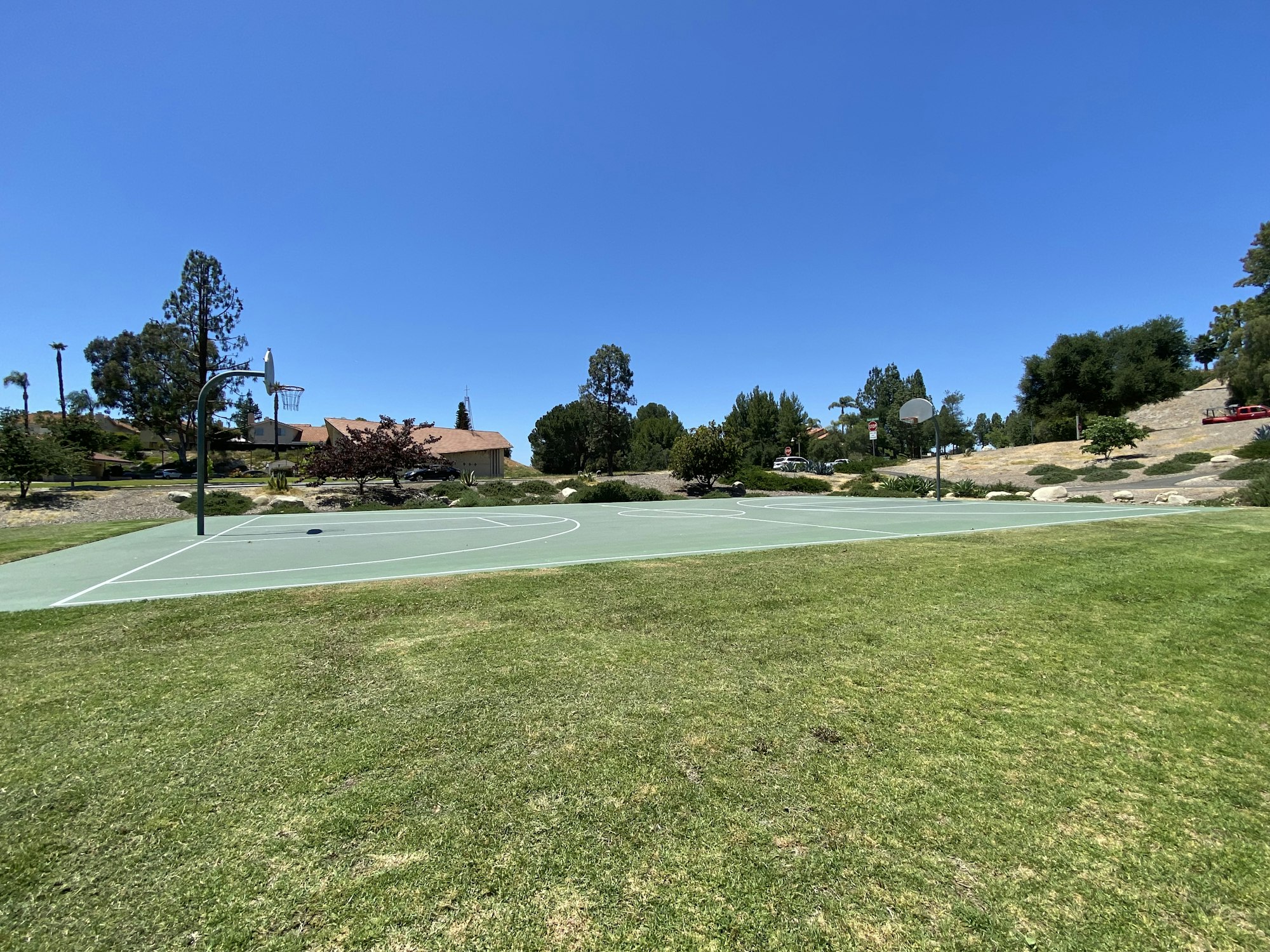An outdoor basketball court surrounded by grass, trees, and houses under a clear blue sky.