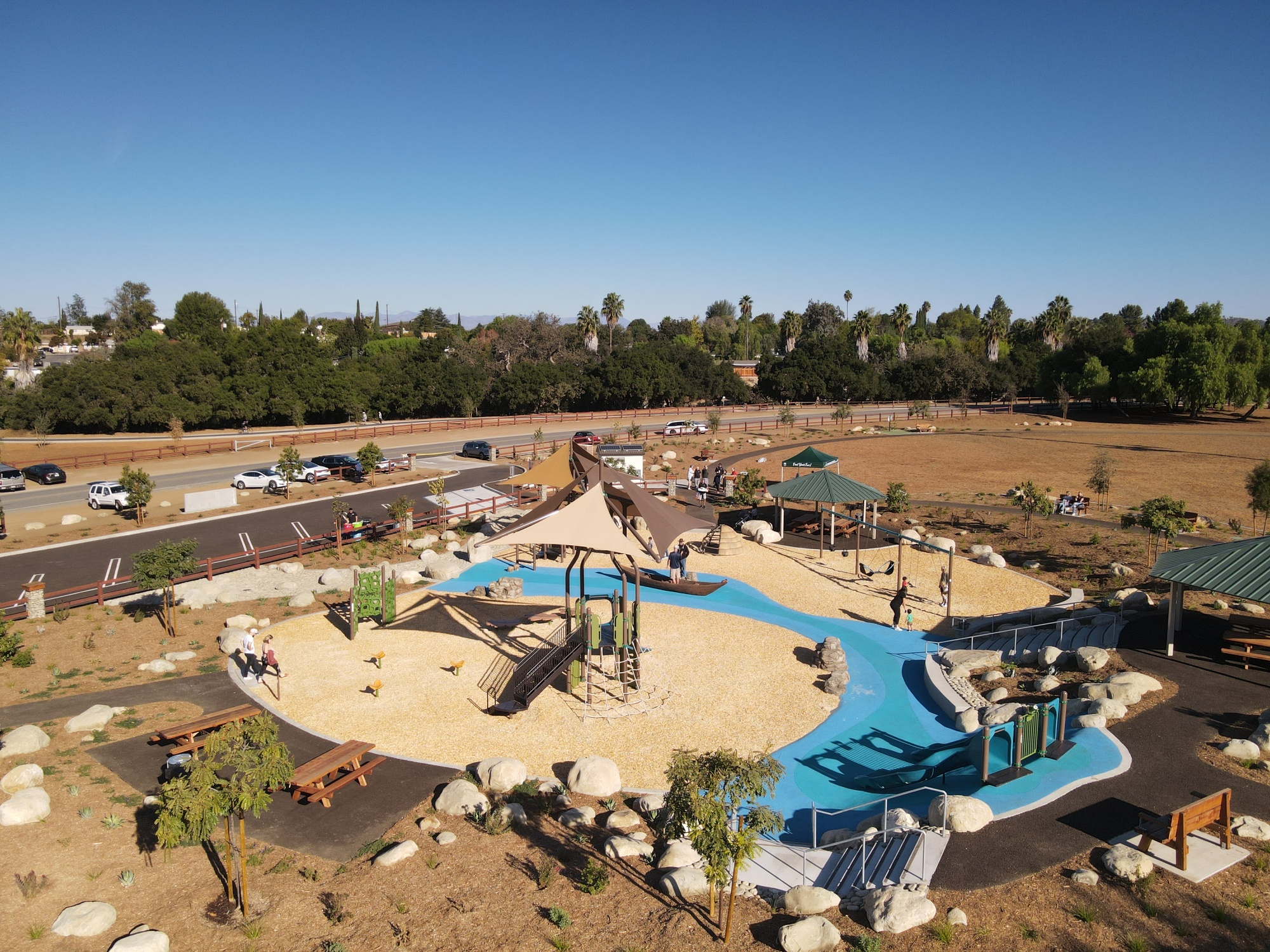 A playground with slides, swings, and shade structures, surrounded by grassy fields and a parking lot. People are enjoying the area.