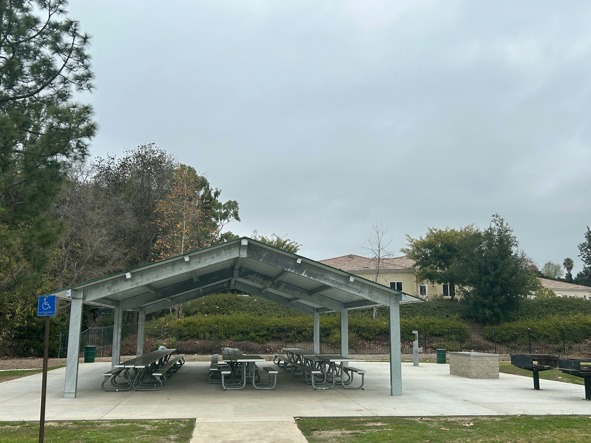 Covered picnic area with tables, trash cans, an accessible parking sign, and a barbecue grill, surrounded by trees and a building in the background.