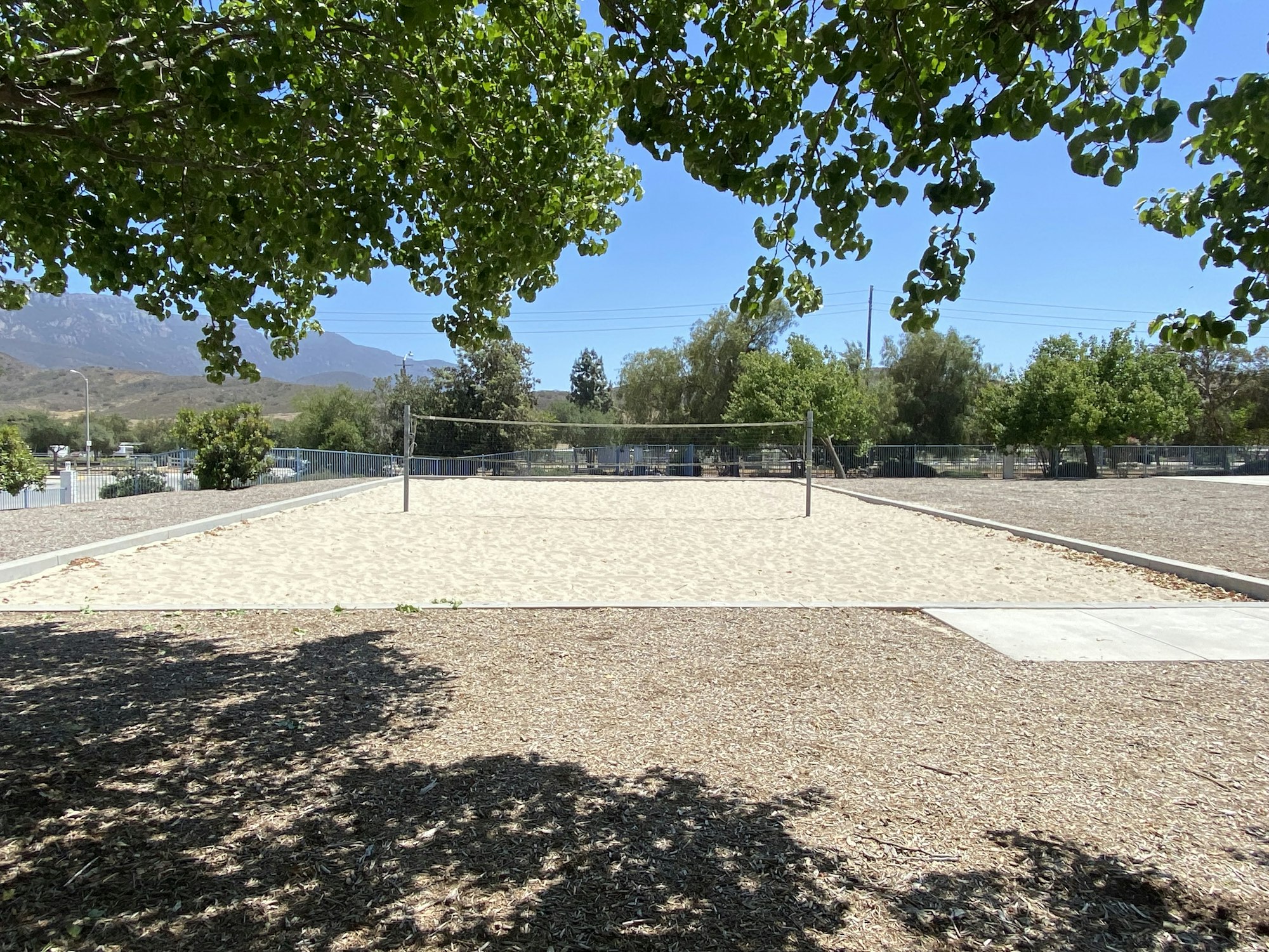 A sandy volleyball court under a clear sky, surrounded by trees and mountains in the background.