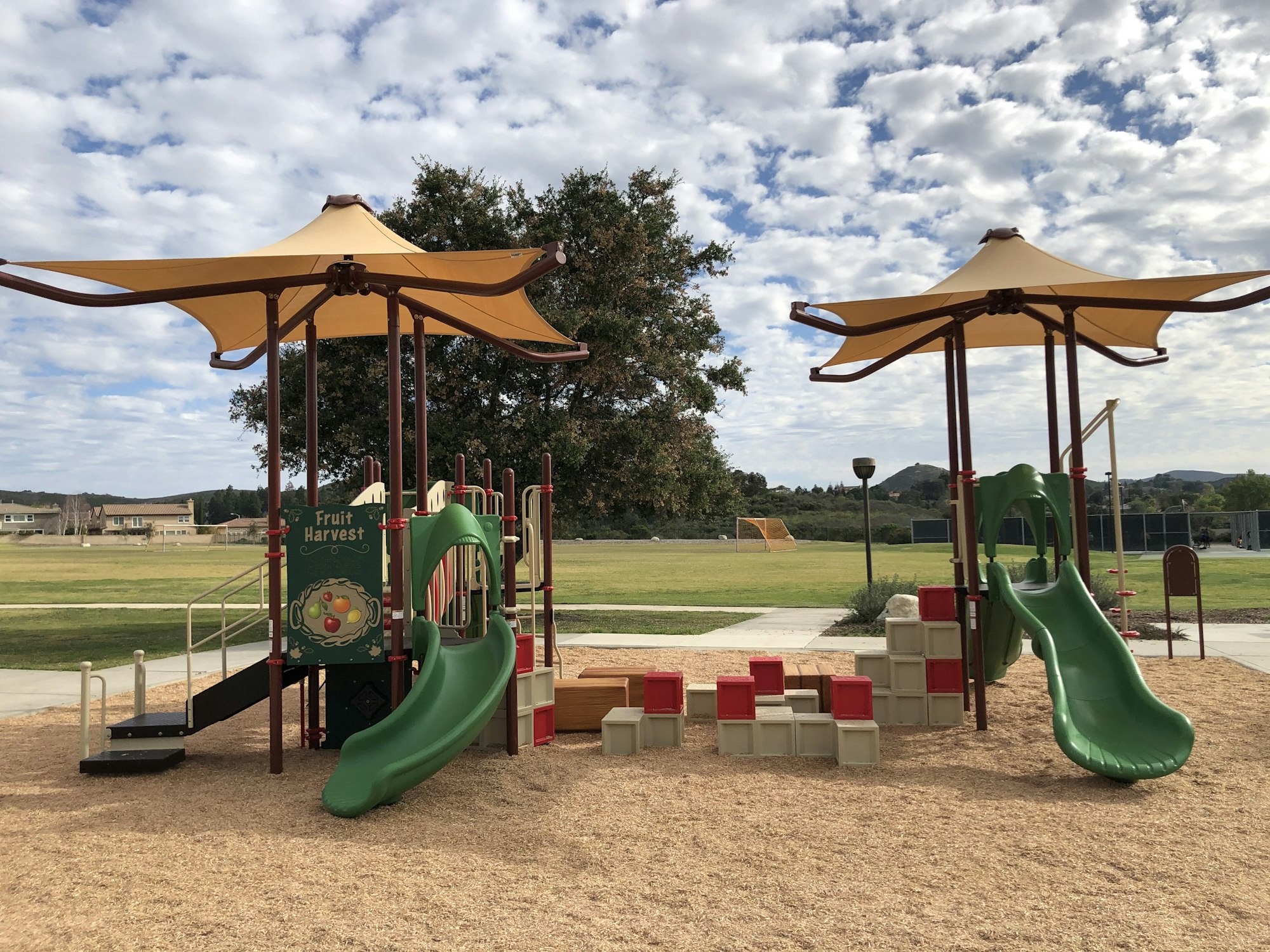 A playground with slides, climbers, and canopy shades, set in a grassy park with trees and a cloudy sky.
