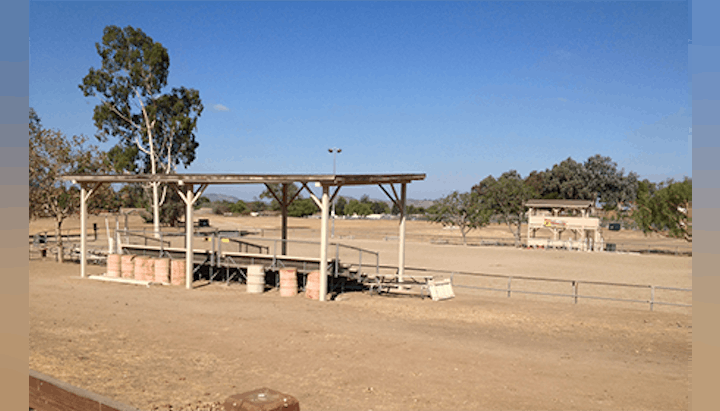 An outdoor shooting range with covered benches, barrels, a distant building, and trees under a clear blue sky.