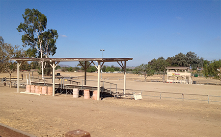 An outdoor shooting range with covered benches, barrels, a distant building, and trees under a clear blue sky.