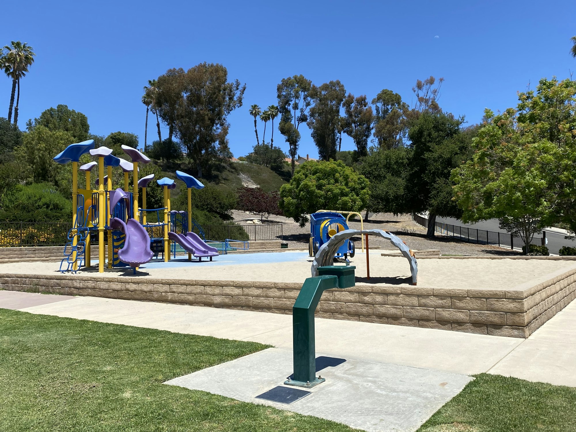 Playground with colorful slides and climbing structures surrounded by trees and grass, under a clear blue sky.