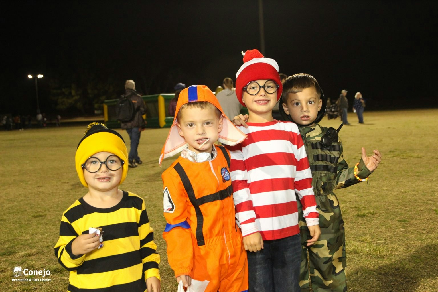 Four kids in costumes: bee, astronaut, striped shirt with hat, and camo outfit, smiling outdoors at night.