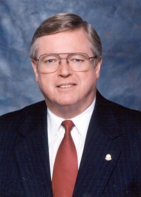 A man in a suit and tie, wearing glasses, with a neutral expression, against a blue-gray background.