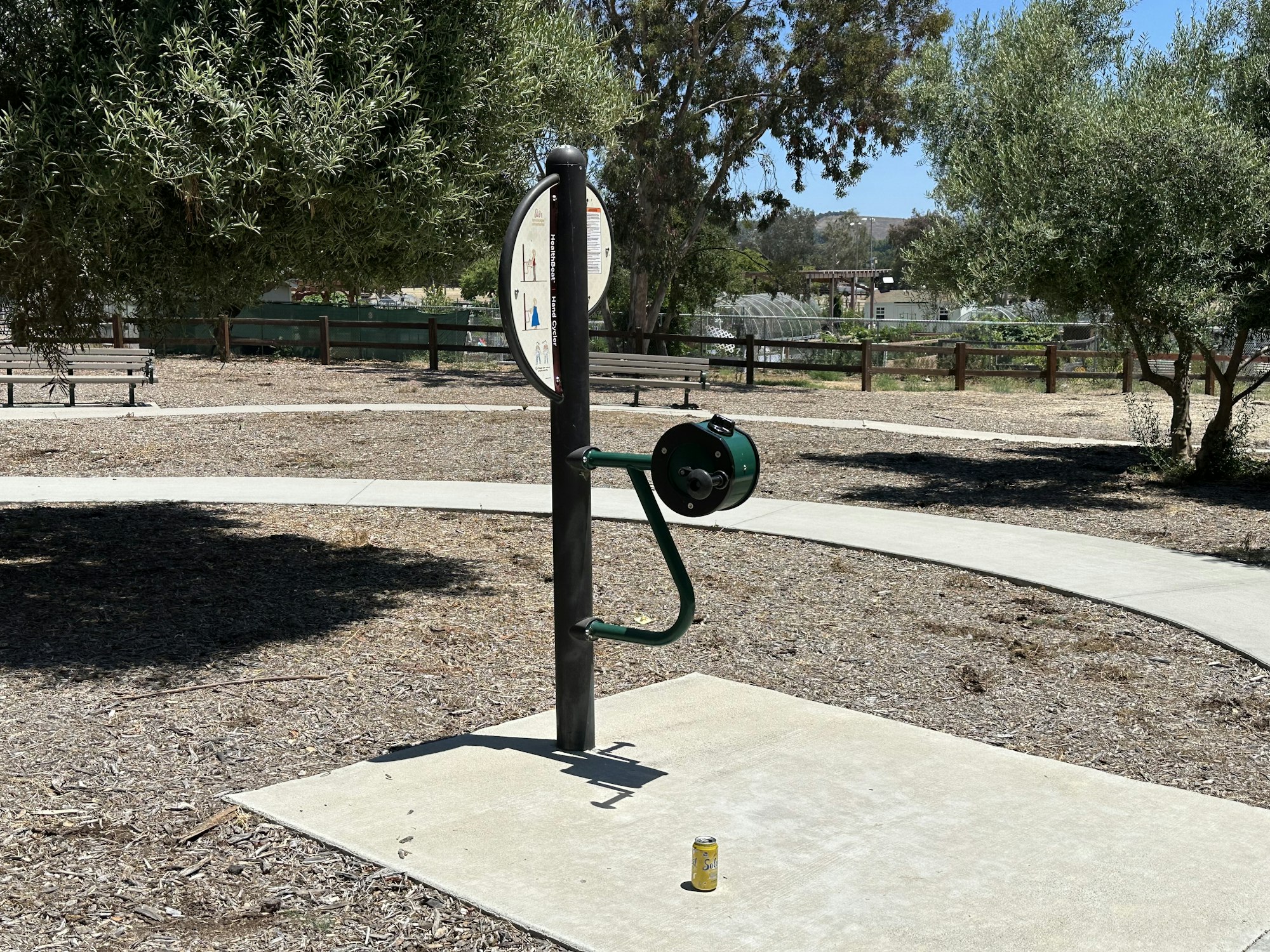 An outdoor fitness station with instructional signage, a green exercise device, and a can on the ground in a park setting.