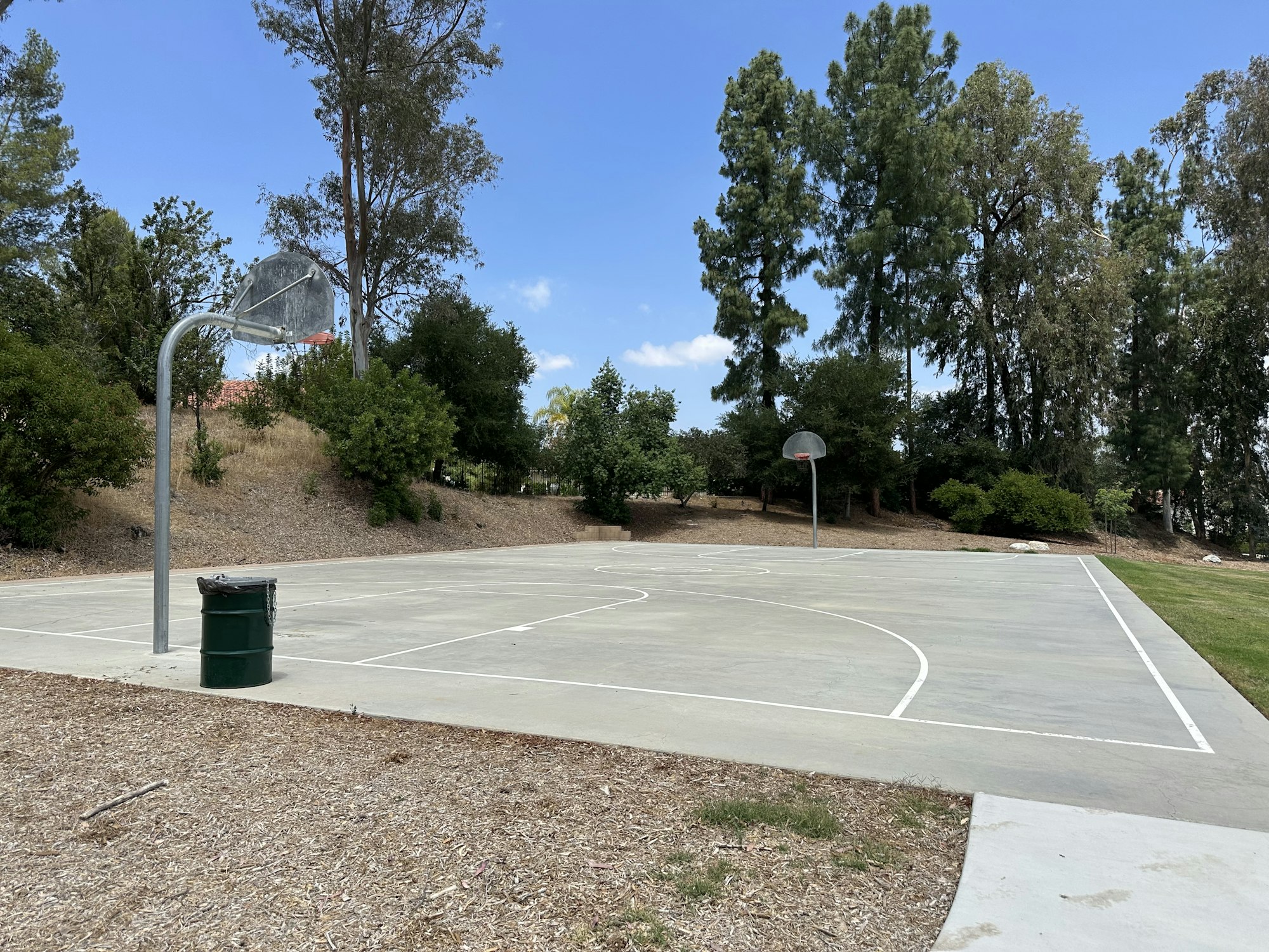 A basketball court surrounded by trees, with two hoops, a trash can, and a clear blue sky in the background.