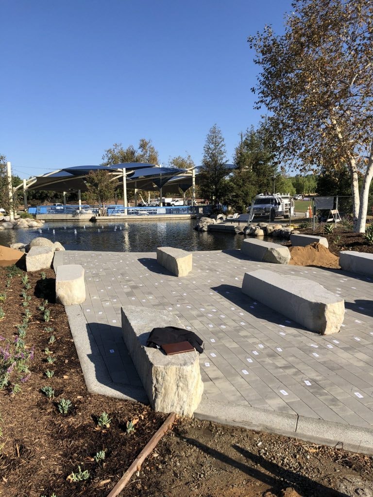 Paved area with large stone benches, some plants, and a pond with shaded structures in the background.