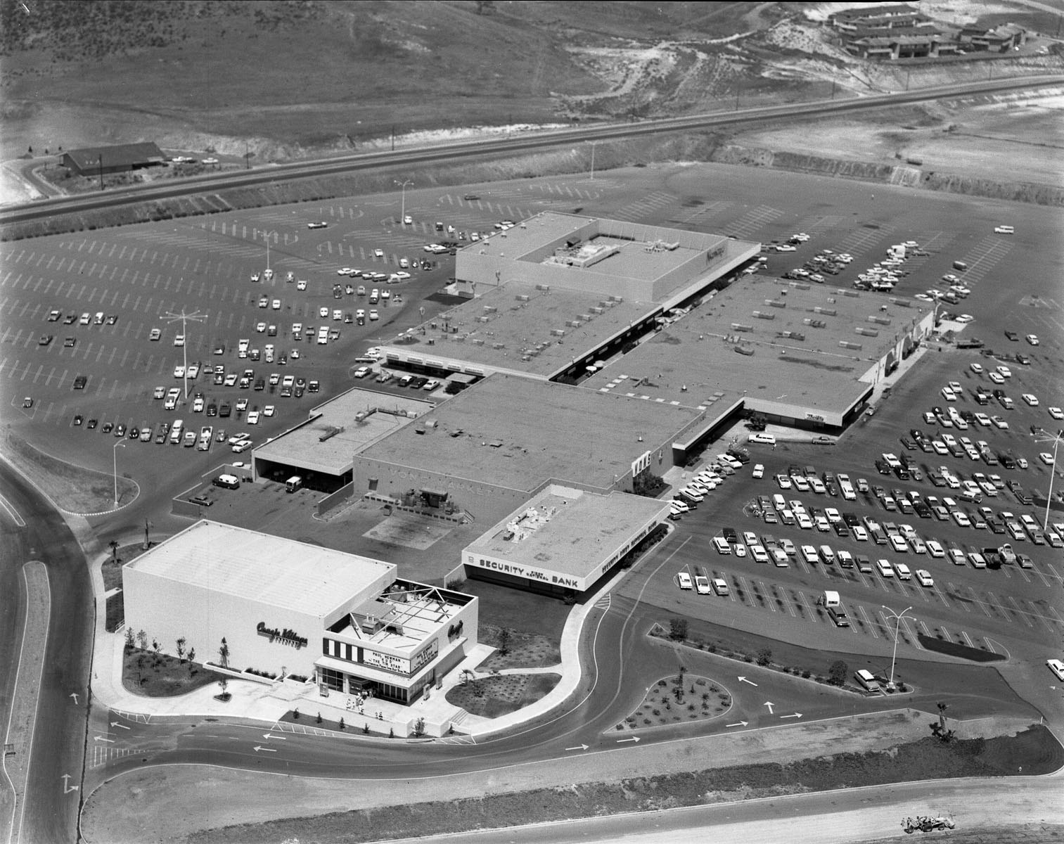 Aerial view of a large 1960s shopping mall with multiple connected buildings, surrounded by a vast parking lot with numerous cars.