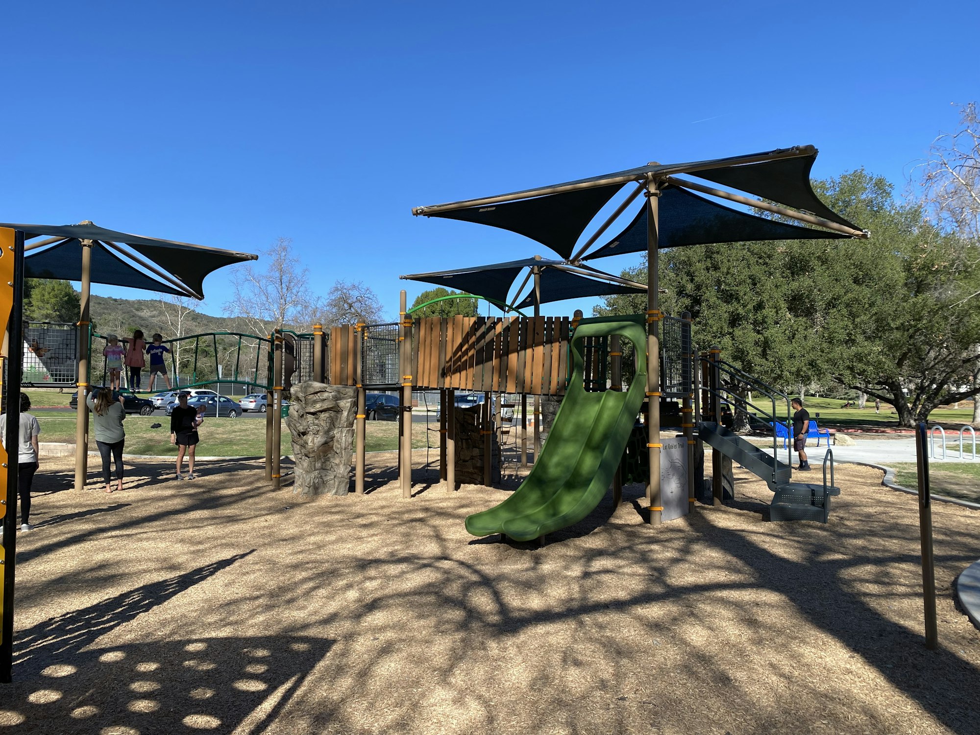 A playground with a slide, climbing structures, and shade covers, surrounded by trees and people enjoying the area.