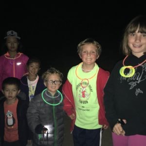 A group of kids in a dark setting, wearing glow sticks around their necks and holding flashlights, smiling at the camera.