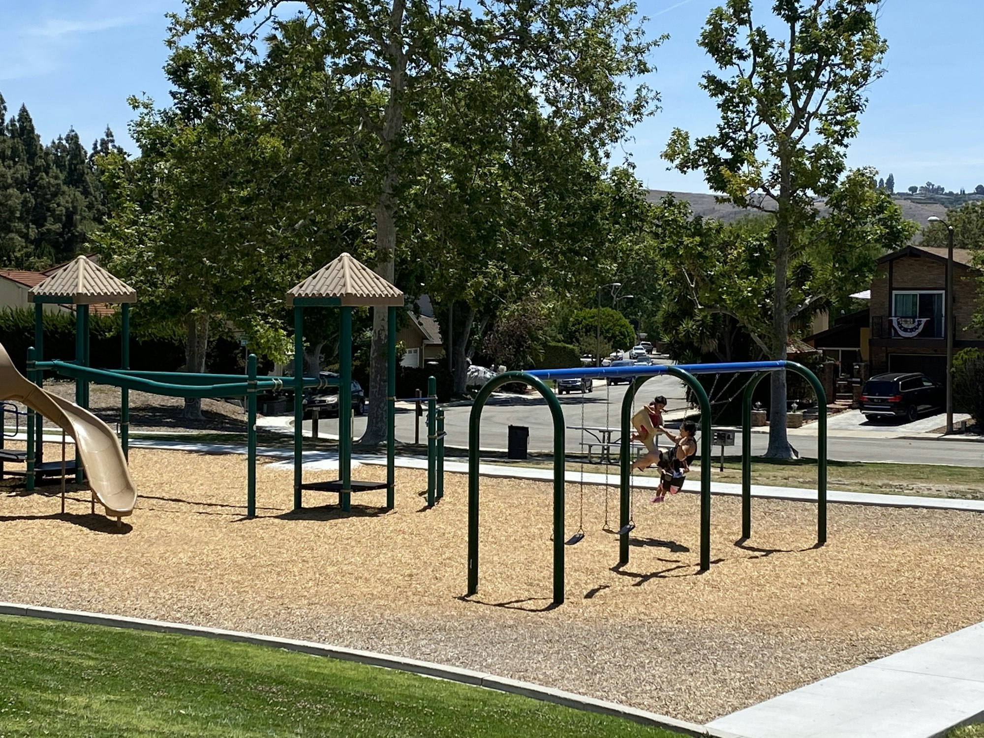 A playground with a slide and swings, surrounded by trees and a residential street.