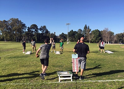 People playing cornhole on a grassy field with trees and a clear blue sky in the background.