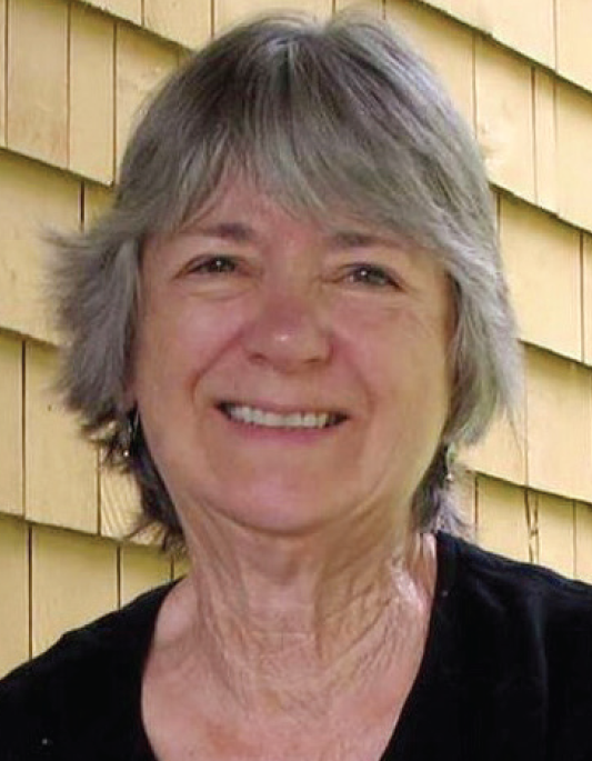 A person with gray hair is smiling, standing in front of a wooden wall.
