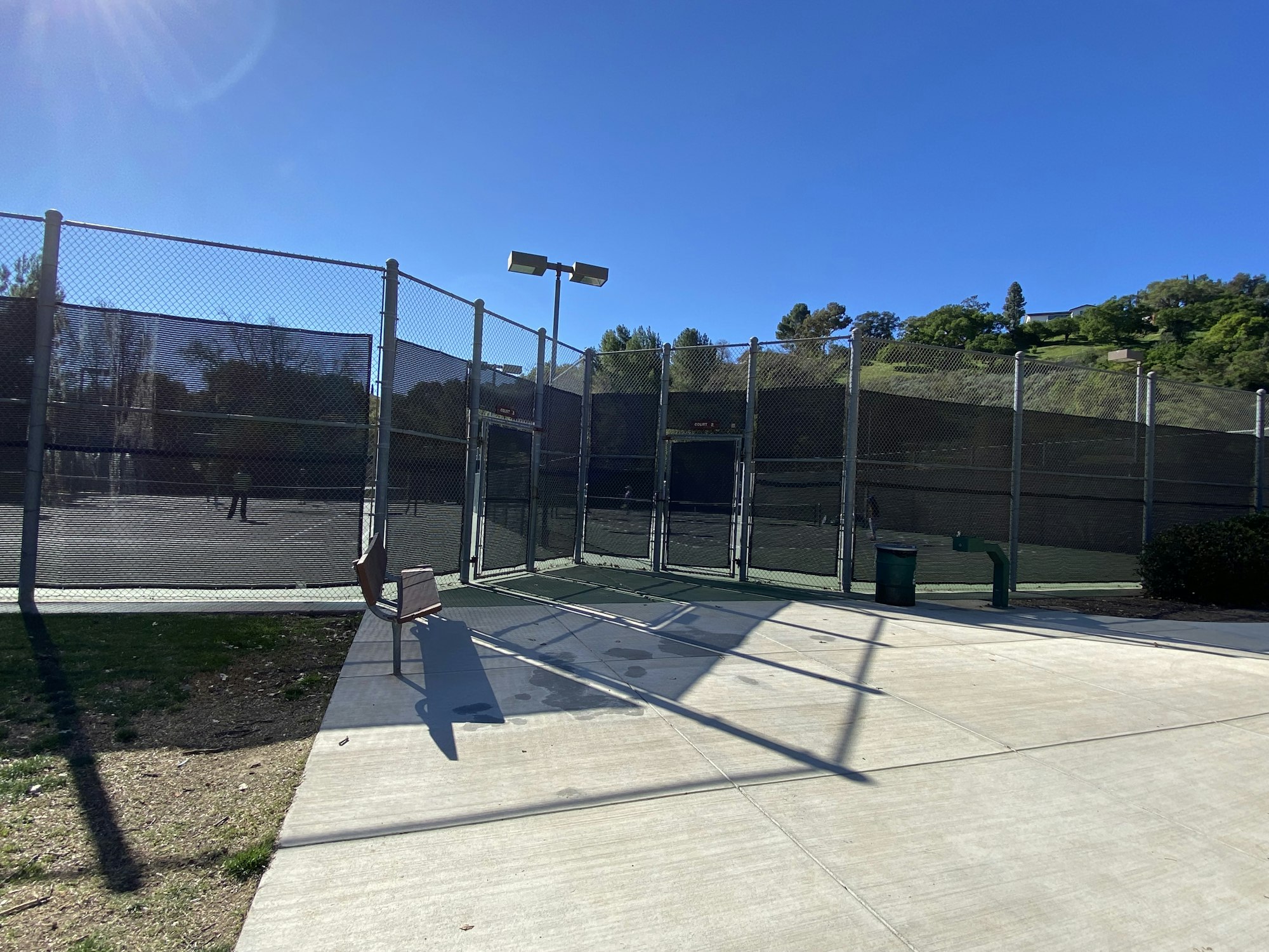 A tennis court with chain-link fencing, a bench, and a trash can under a clear blue sky.