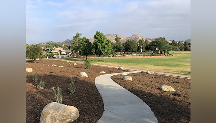 A park with a winding path, grass, scattered rocks, trees, a playground, and hills in the background under a cloudy sky.