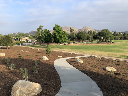 A park with a winding path, grass, scattered rocks, trees, a playground, and hills in the background under a cloudy sky.