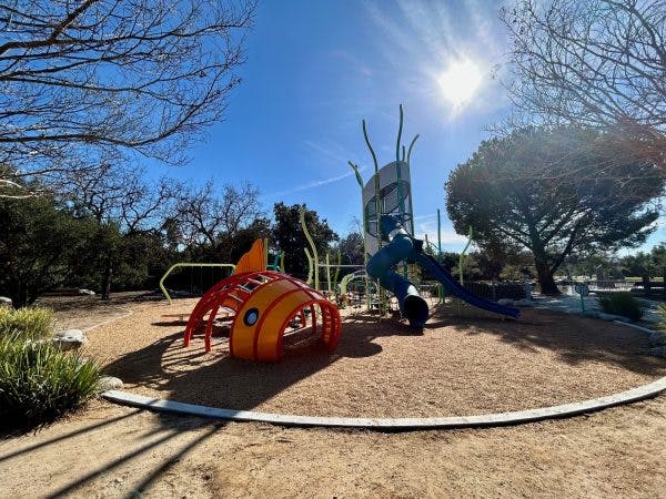 A playground with colorful climbing structures and slides under a sunny sky, surrounded by trees.