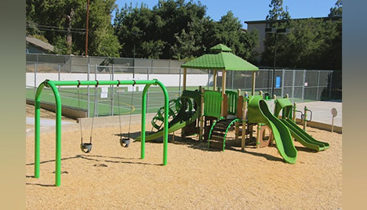 Playground with green slides, swings, and climbing structure on a sandy surface, near a fenced tennis court.