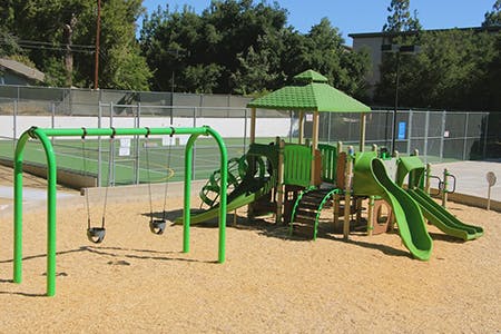 Playground with green slides, swings, and climbing structure on a sandy surface, near a fenced tennis court.