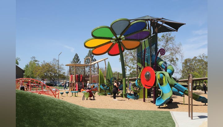 Colorful playground with a giant flower structure, slides, climbing nets, and people enjoying the sunny day.