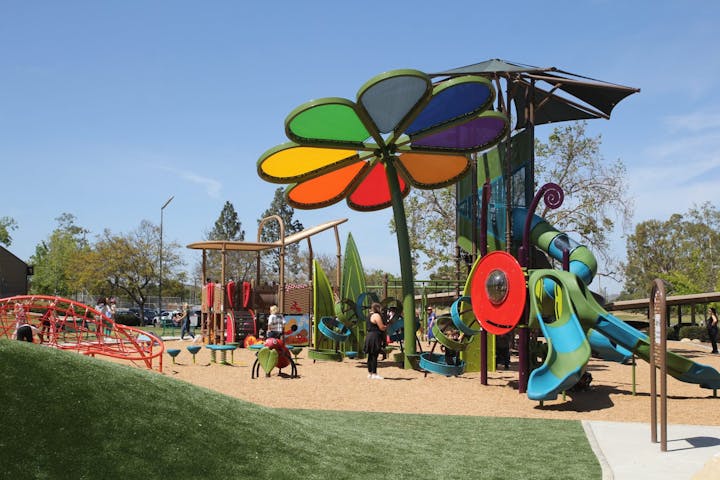 Colorful playground with a giant flower structure, slides, climbing nets, and people enjoying the sunny day.