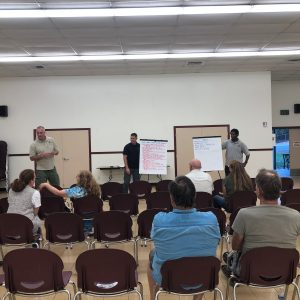 A group of people in a room with three speakers in front, whiteboards with notes, and attendees sitting in chairs.