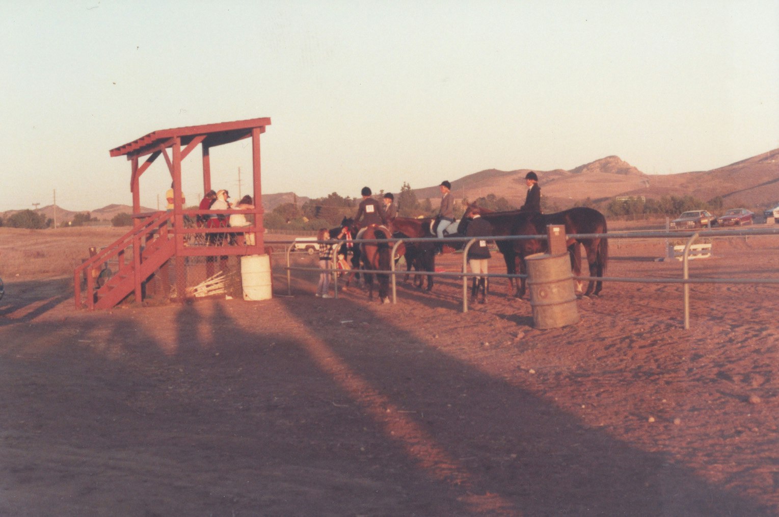 A group of people watching horseback riders near a red wooden platform, set in a rural landscape.