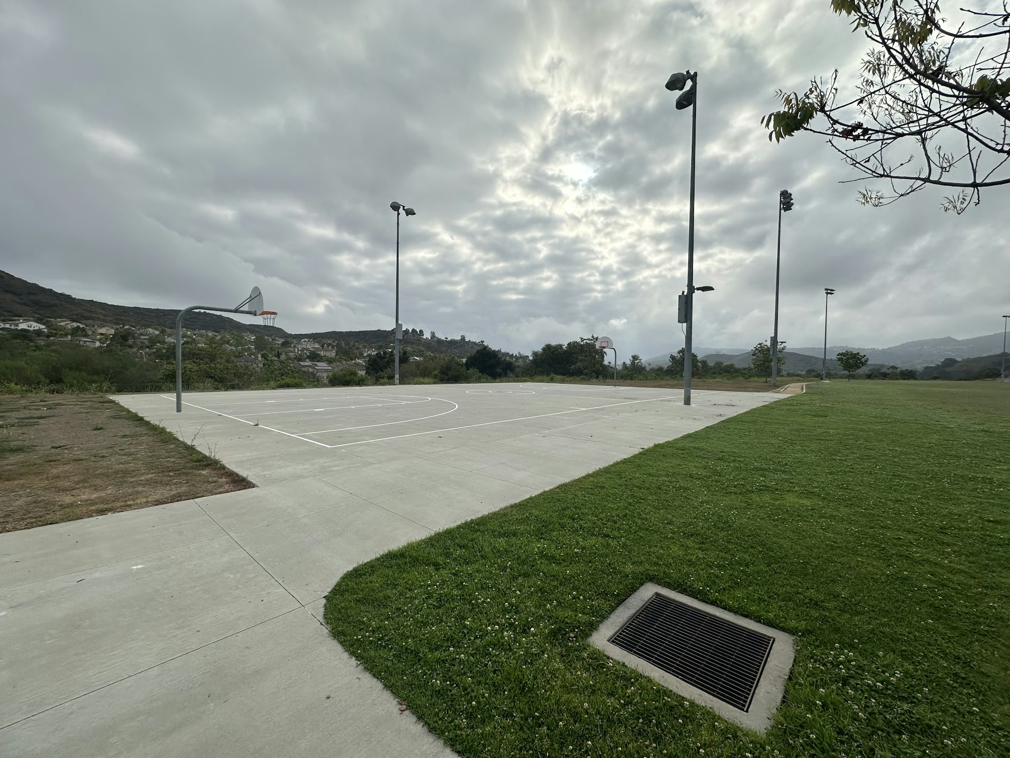 An empty outdoor basketball court with two hoops, surrounded by grassy areas and a cloudy sky above.