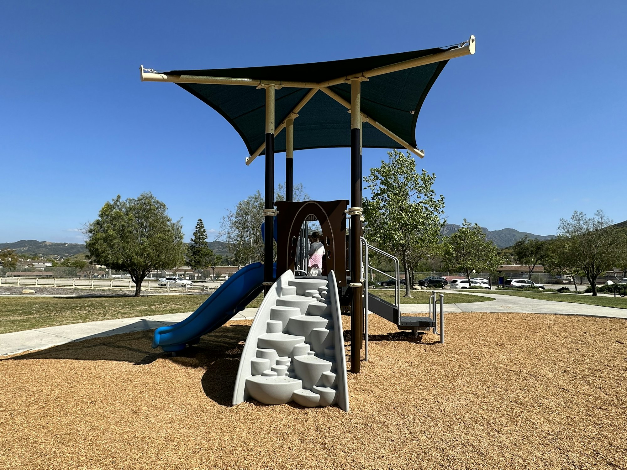 Playground with a slide, climbing steps, and shade. Trees and mountains in the background under a clear blue sky.