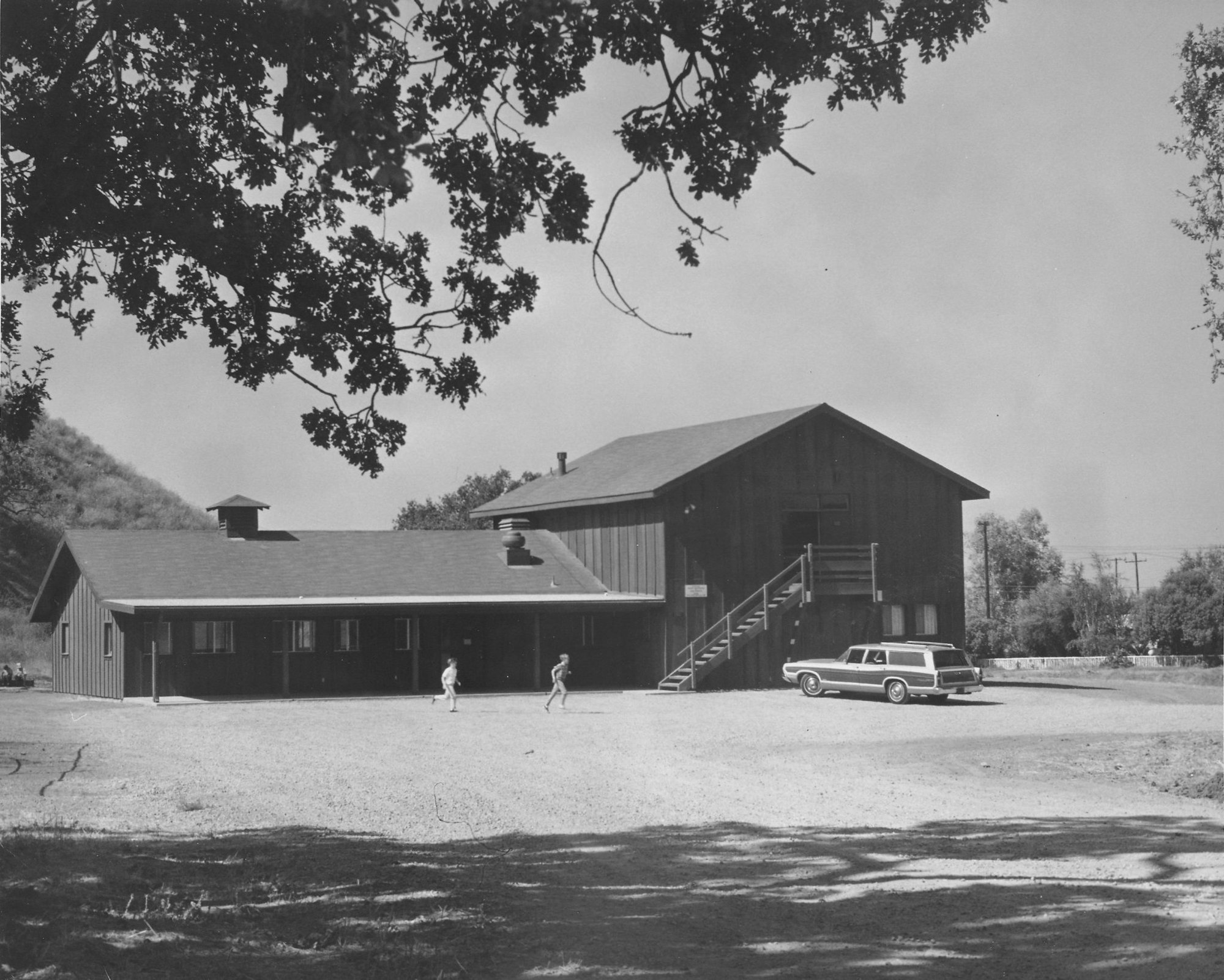 A barn-like structure with two people running in front, surrounded by trees, and a vintage car parked nearby.