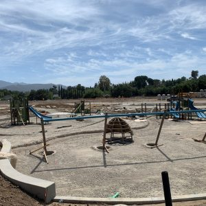 A playground under construction with swings and play structures, surrounded by dirt, under a partly cloudy sky.