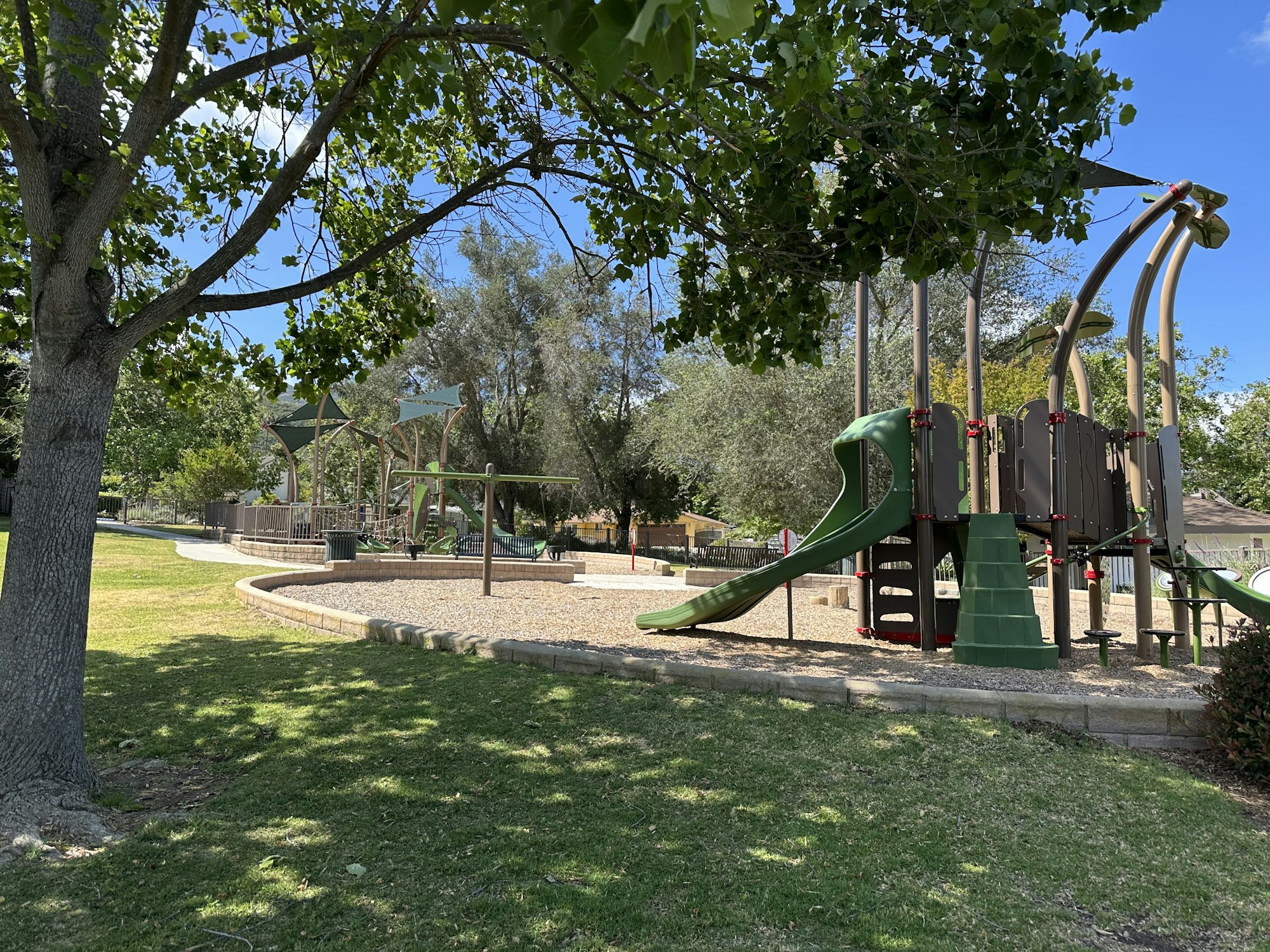 A playground featuring slides and swings, surrounded by trees and green grass on a sunny day.