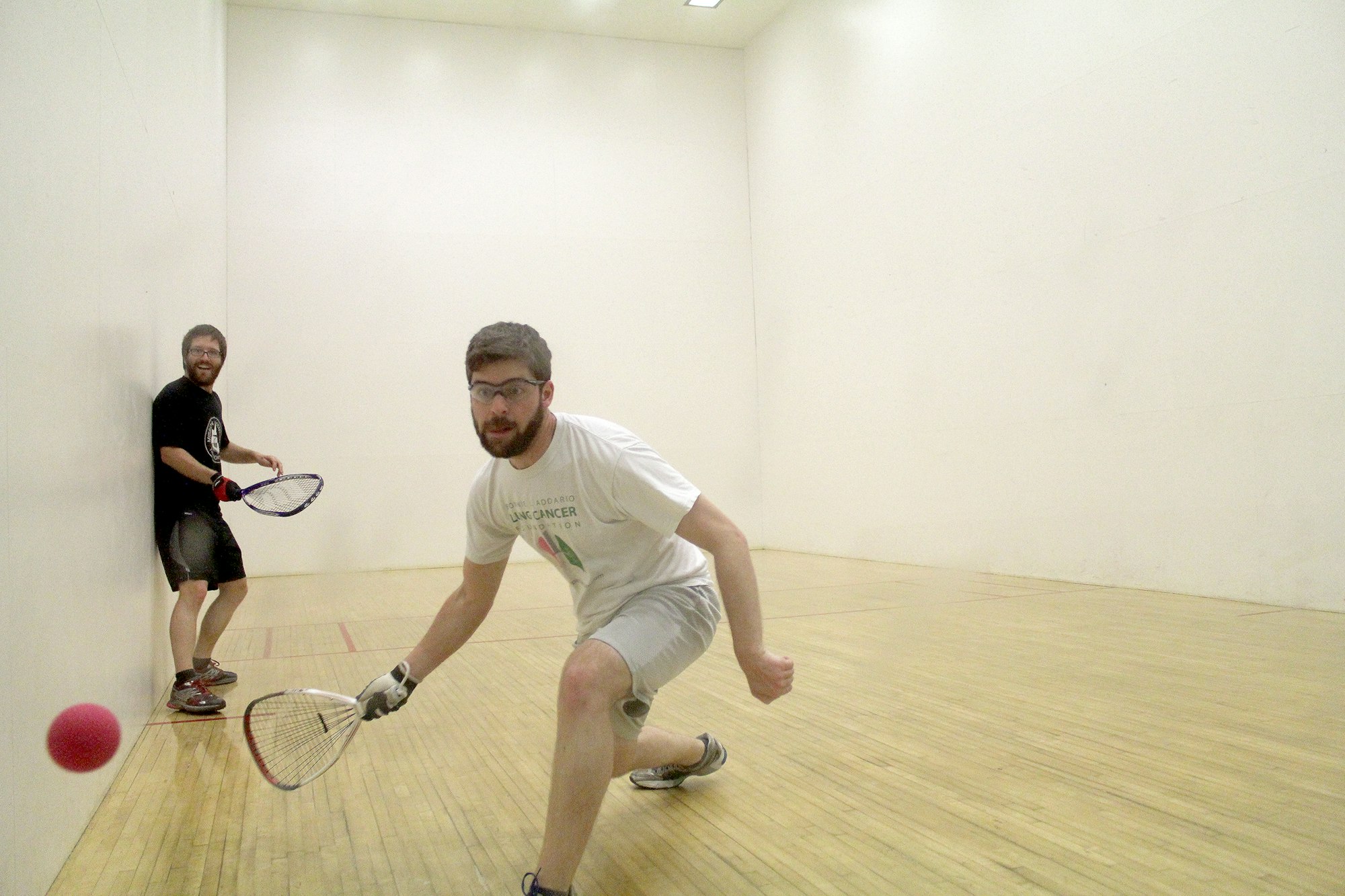 Two people playing racquetball in a court, one hitting a red ball, both wearing protective eyewear and smiling.