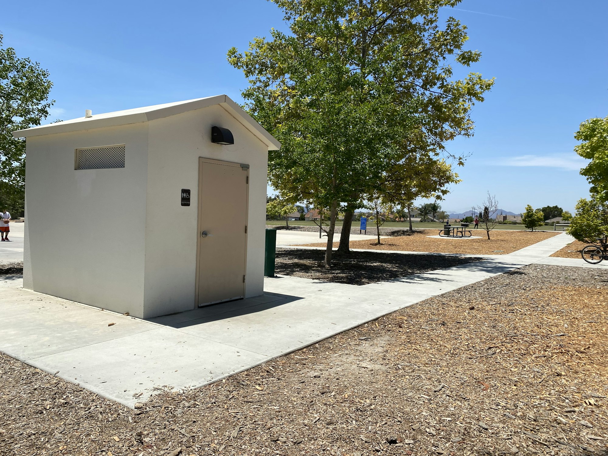 A small restroom building in a park with trees, pathways, and a picnic table in the background.
