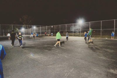 People playing a game on a fenced court at night under bright lights.