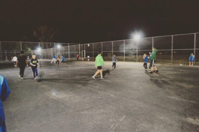People playing a game on a fenced court at night under bright lights.