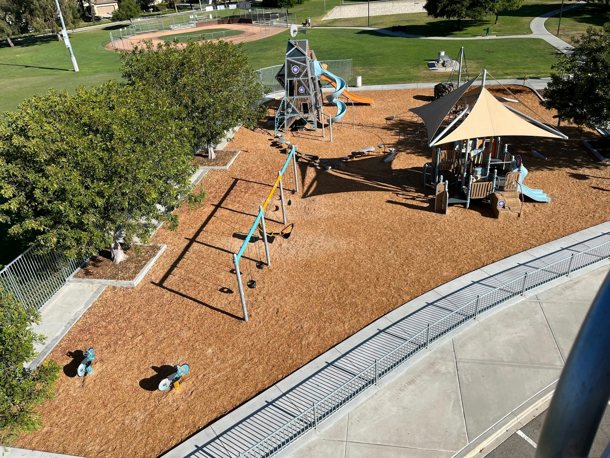 Playground with swings, slides, climbing structure, and trees, near a baseball field.