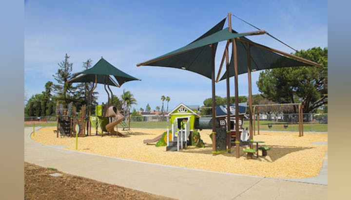 A playground with climbing structures, slides, shade canopies, and a playhouse, surrounded by trees and a clear sky.