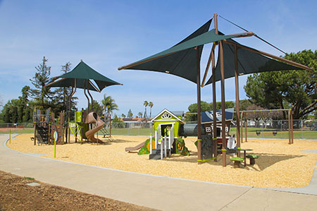 A playground with climbing structures, slides, shade canopies, and a playhouse, surrounded by trees and a clear sky.